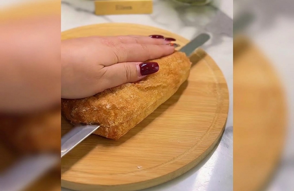 Dłoń trzymająca złotą ciabattę, podczas gdy nóż z ząbkami przekrawa ją na pół na drewnianej desce.