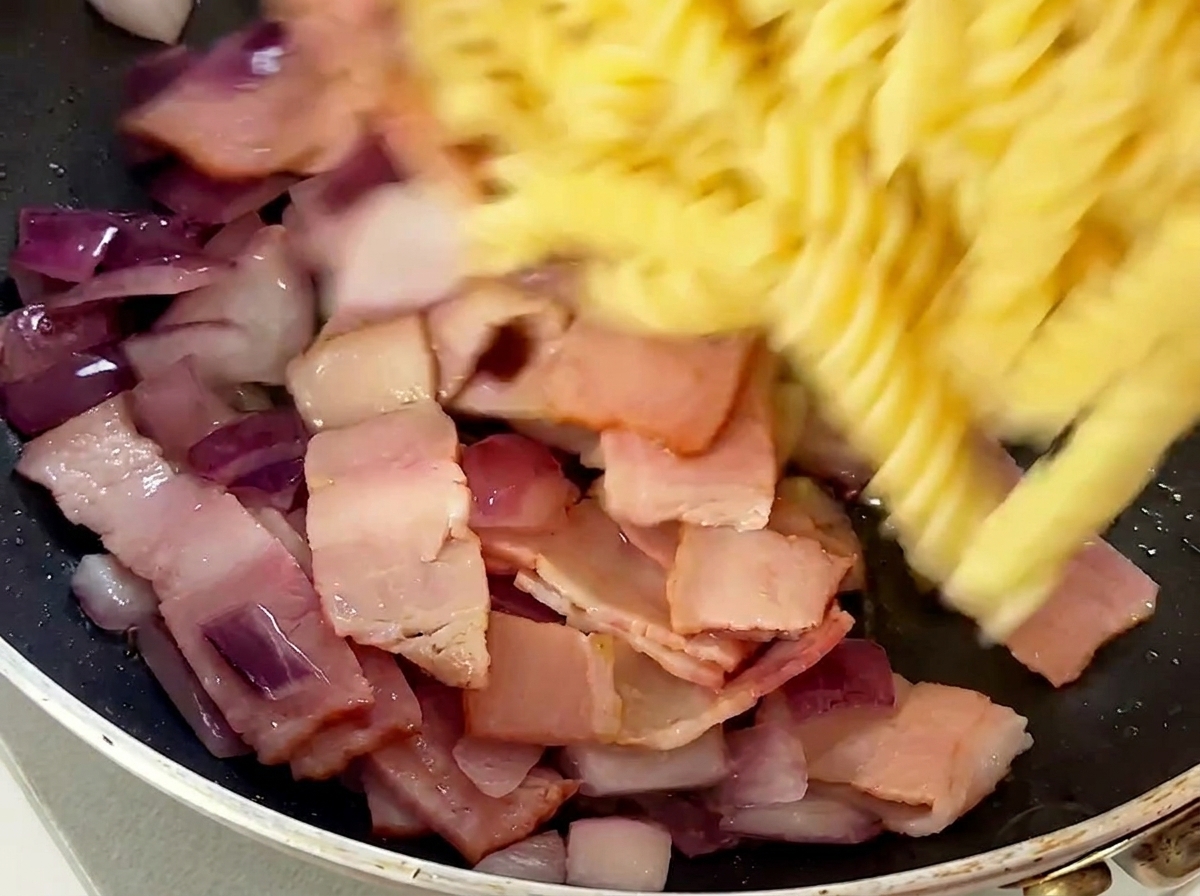 Ugotowany makaron fusilli dodawany do patelni z boczkiem i cebulą.