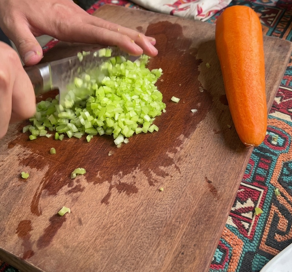 Drobno posiekany seler i marchew na drewnianej desce do krojenia obok całej marchewki.