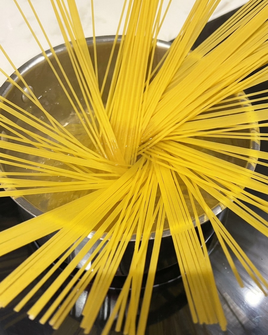 Nitki spaghetti gotujące się w dużym garnku z wodą, widać unoszącą się parę.