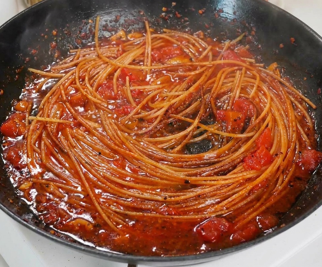 Zbliżenie na ugotowane spaghetti wymieszane w bogatym, gęstym sosie pomidorowym na czarnej patelni.