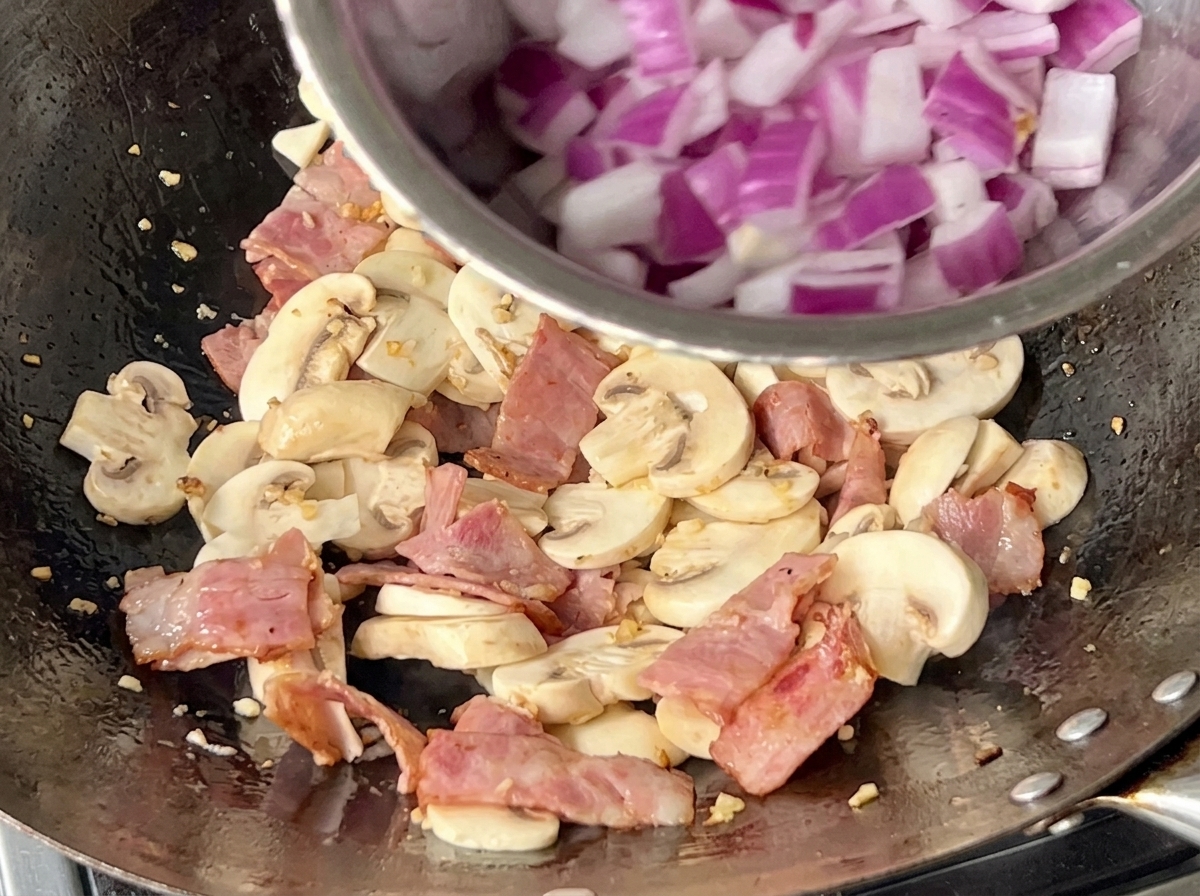 Wok wypełniony pieczarkami, kawałkami boczku i świeżo dodaną czerwoną cebulą.