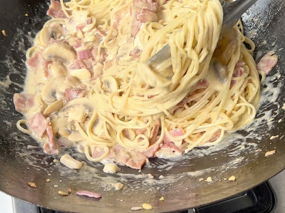 Zbliżenie spaghetti mieszanego w kremowym sosie za pomocą metalowych szczypiec.
