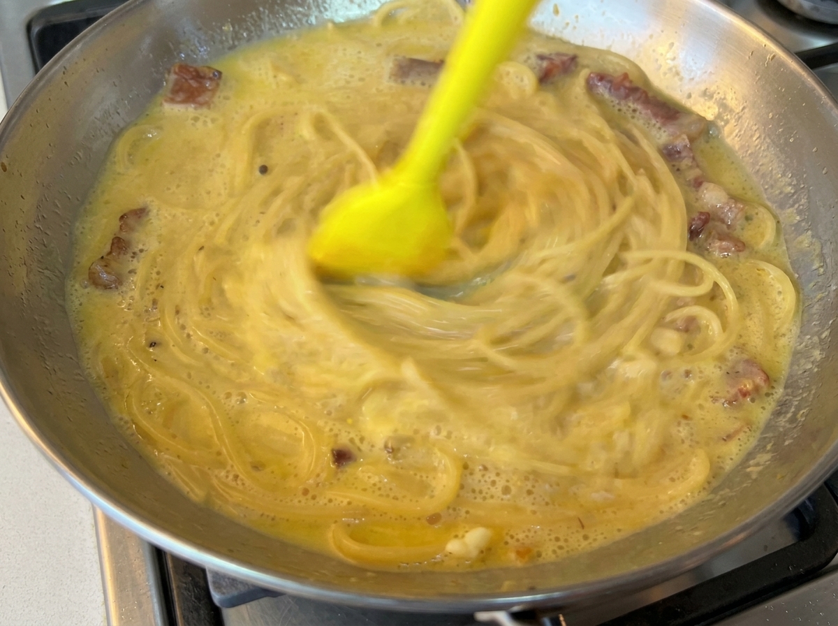 Gumowa szpatułka energicznie mieszająca spaghetti z kremowym sosem na stalowej patelni.