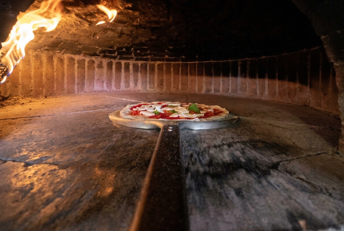 Metalowa łopata przenosi przygotowaną pizzę do gorącego pieca opalanego drewnem.