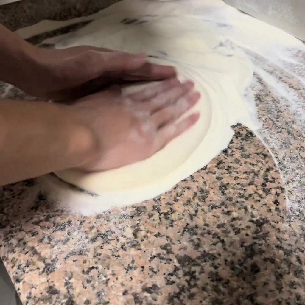 Dłonie delikatnie ugniatają i rozciągają okrągłe ciasto na pizzę na oprószonym mąką blacie.