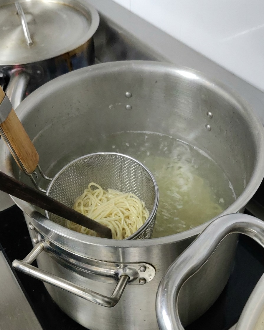 Makaron ramen gotujący się w metalowym sitku zanurzonym w dużym garnku z wodą.