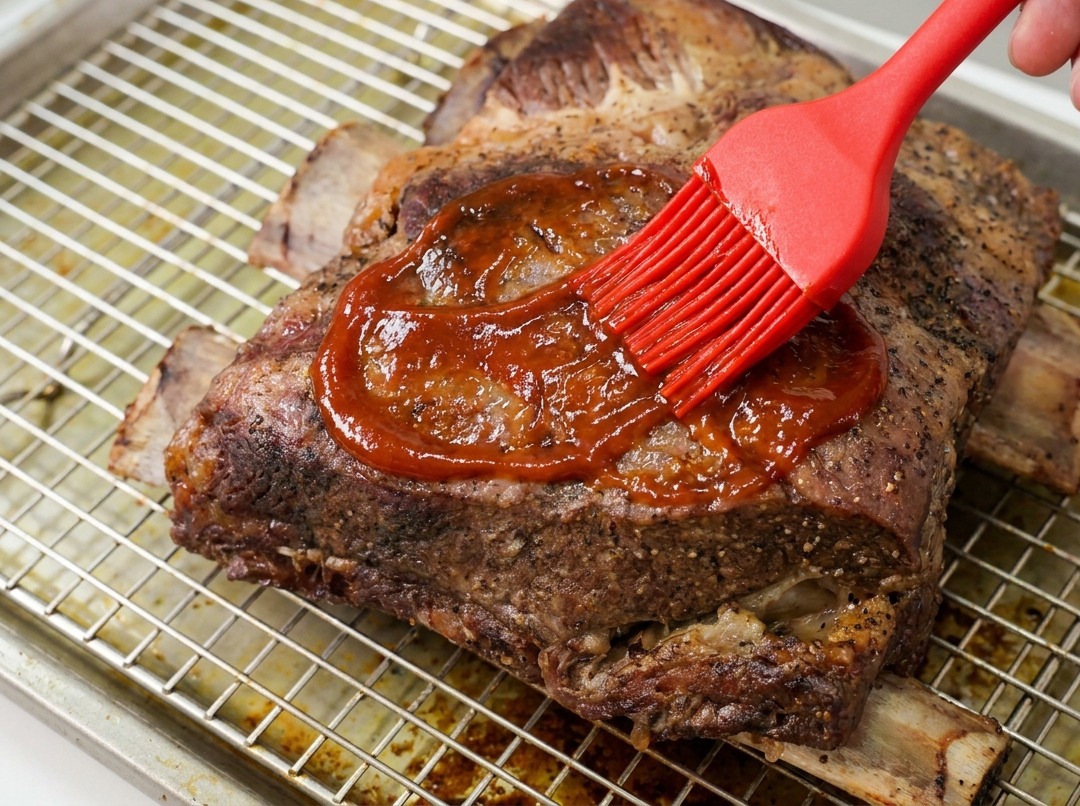 Ręka smarująca pędzlem silikonowym gęsty sos BBQ na upieczonych żeberkach wołowych.