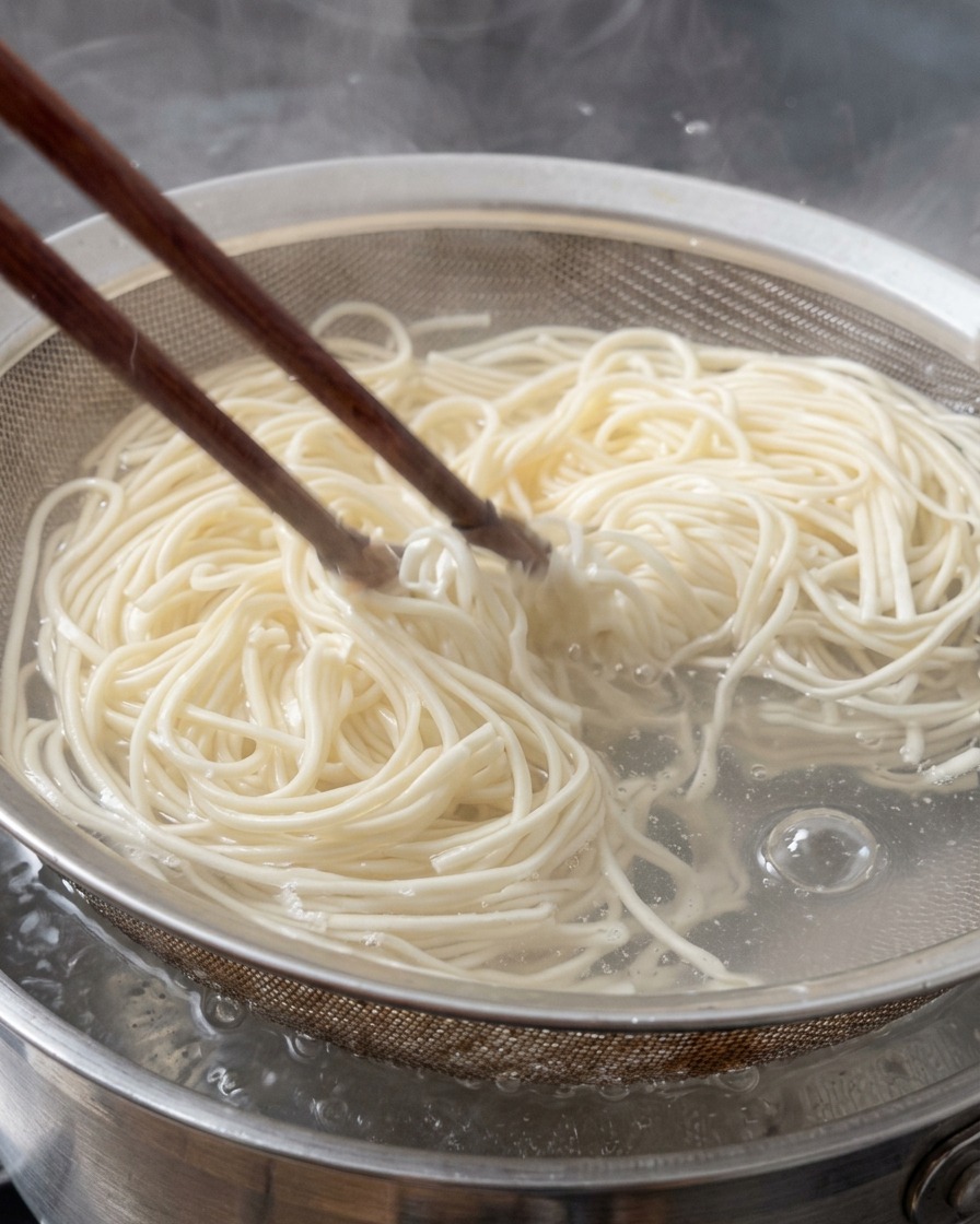 Świeży makaron ramen wyjmowany z wrzątku za pomocą metalowego sitka i pałeczek.