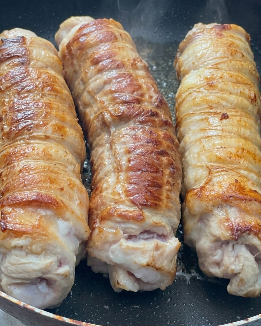 Trzy roladki boczku wieprzowego smażące się na patelni, z widoczną złocistą skórką.
