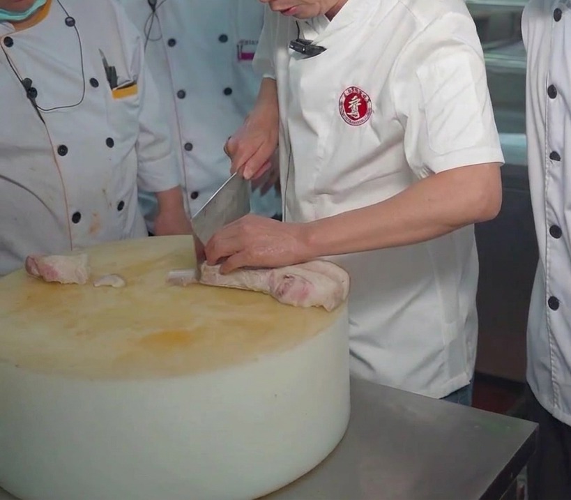 Szef kuchni krojący tasakiem kawałek gotowanego boczku na dużej desce do krojenia.