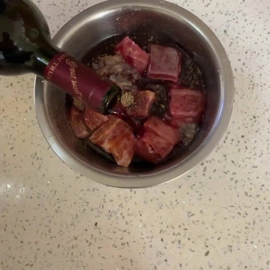 Czerwone wino przelewane z ciemnej butelki na przyprawiony boczek w stalowej misce.