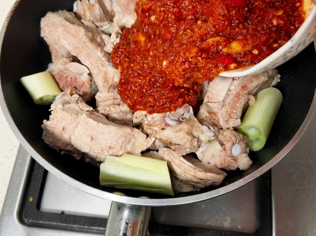 Gęsta, czerwona pasta z chili wylewana z miski na zblanszowane żeberka na patelni.