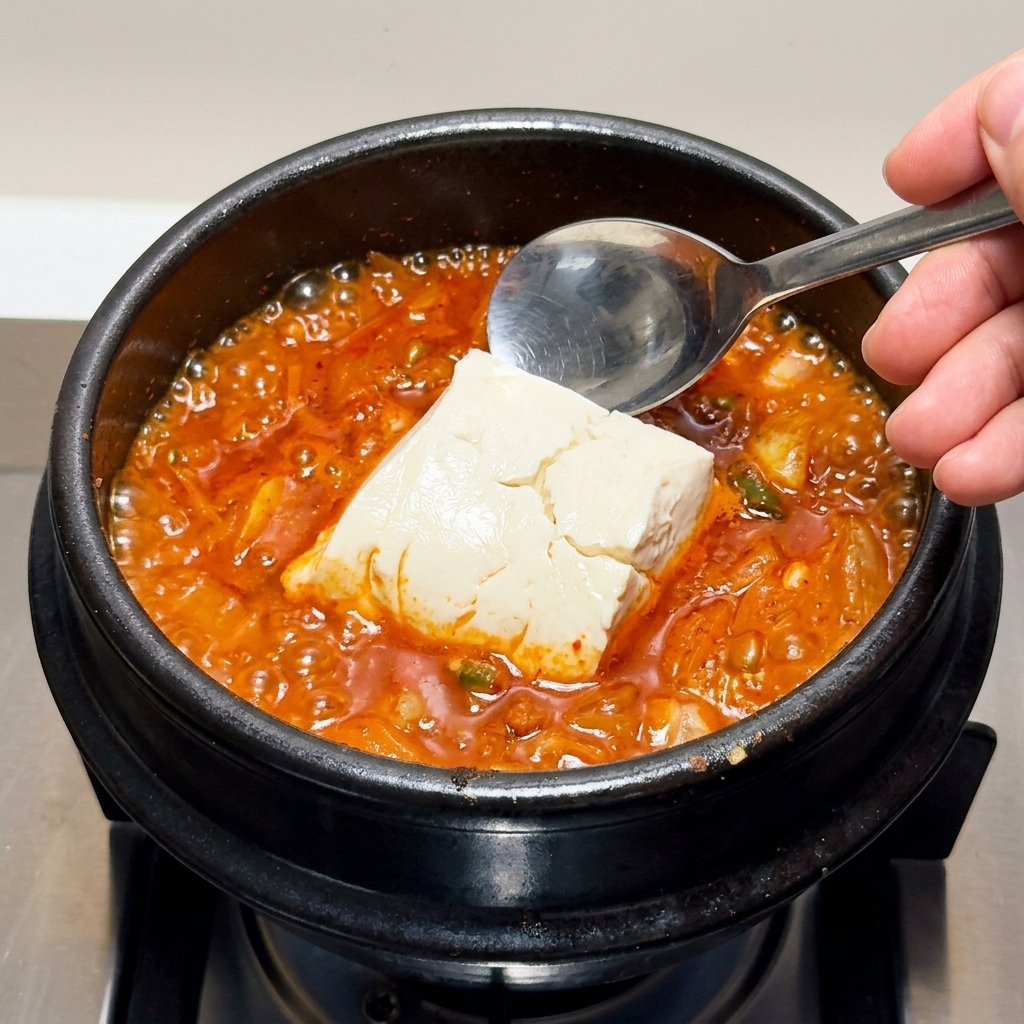Duże kawałki jedwabistego tofu ostrożnie nakładane łyżką do gotującego się gulaszu kimchi.