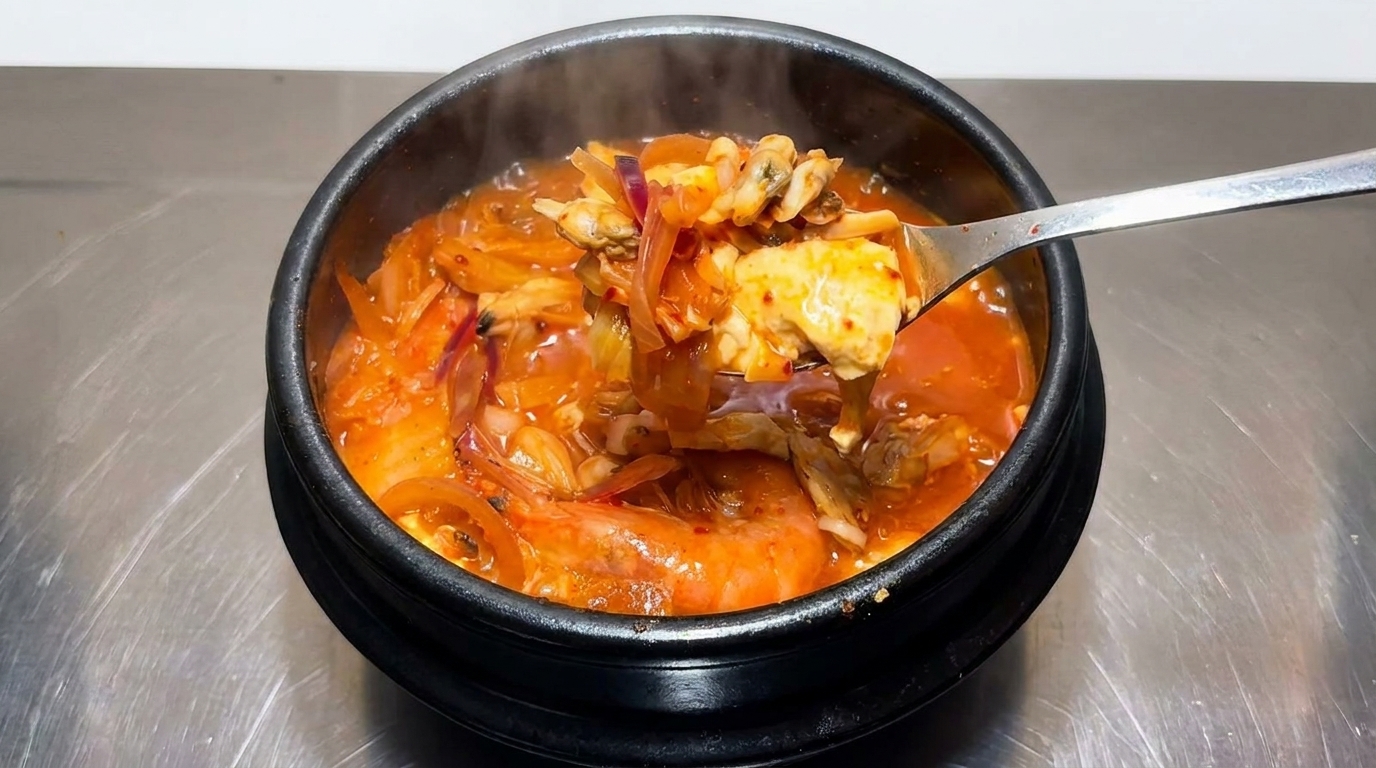 Łyżka pełna bogatego, czerwonego gulaszu kimchi z małżami, krewetkami, tofu i cebulą, wyjęta prosto z czarnego kamiennego garnka.