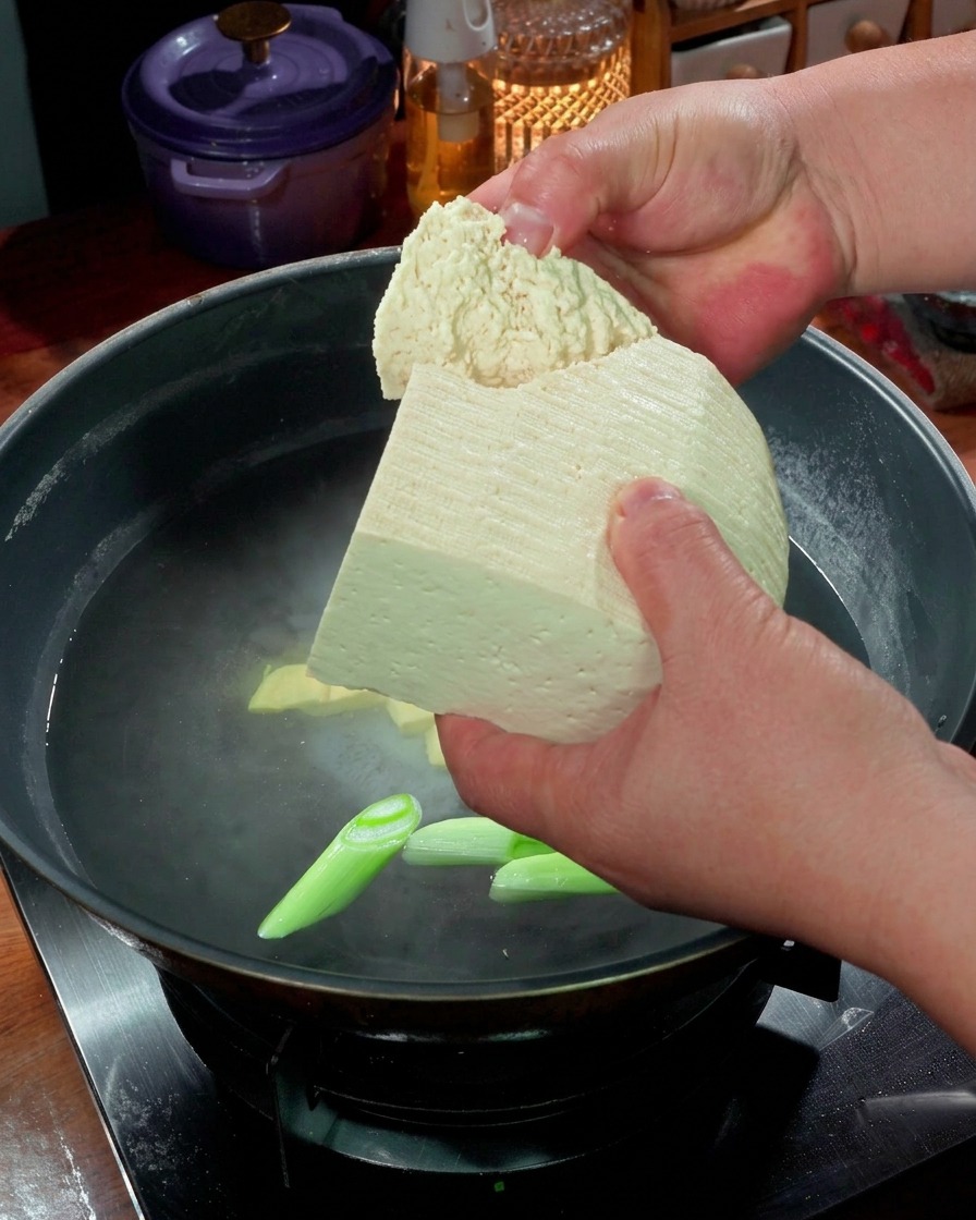 Ręce łamiące duże kawałki twardego tofu do garnka z wrzącą wodą i zieloną cebulką.