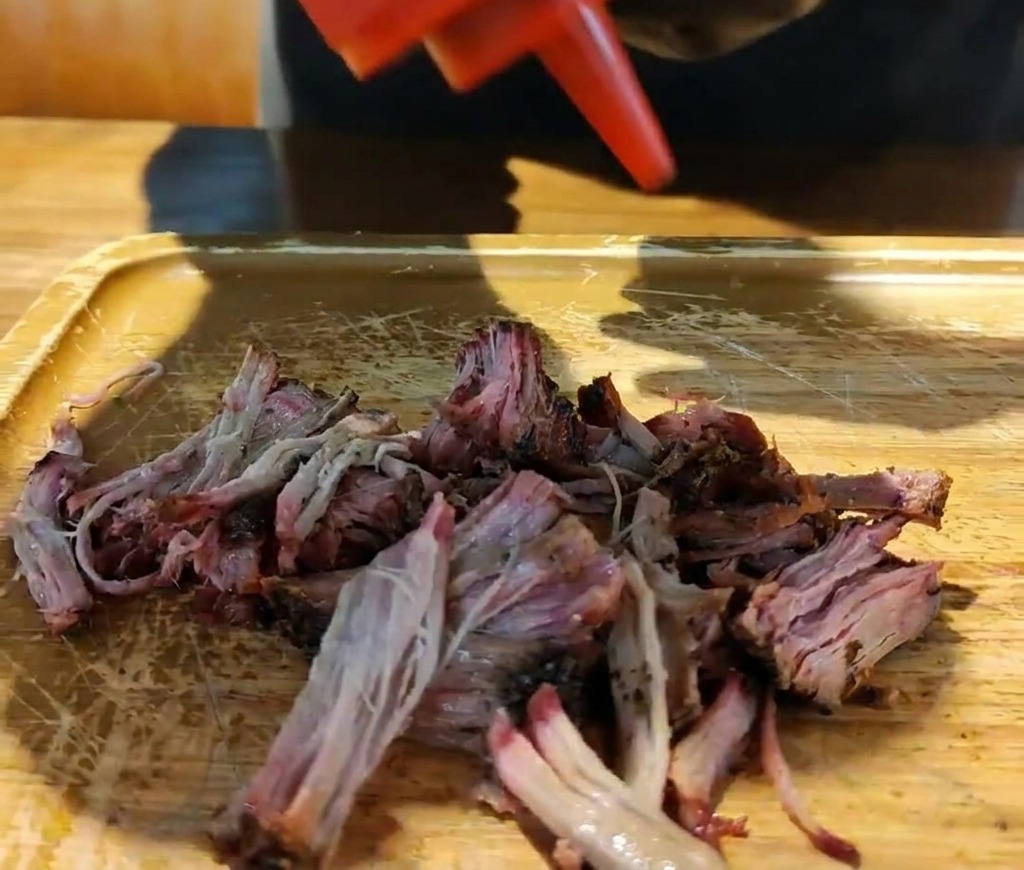 Czerwona butelka, z której gęsty sos barbecue spływa na stertę rozdrobnionej wędzonej wieprzowiny na desce.