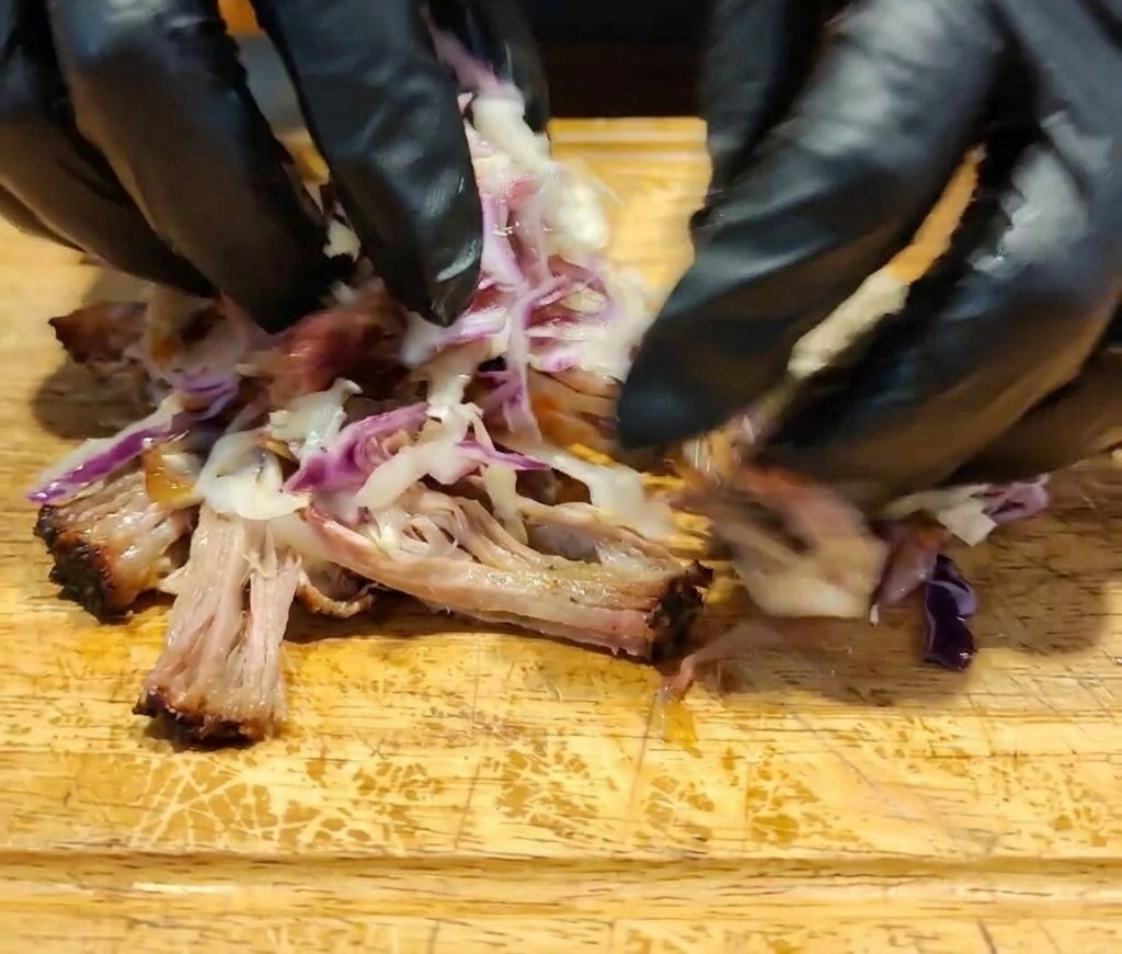 Dłonie w rękawiczkach mieszające rozdrobnione mięso, ciemny sos i kolorowy coleslaw na drewnianej powierzchni.