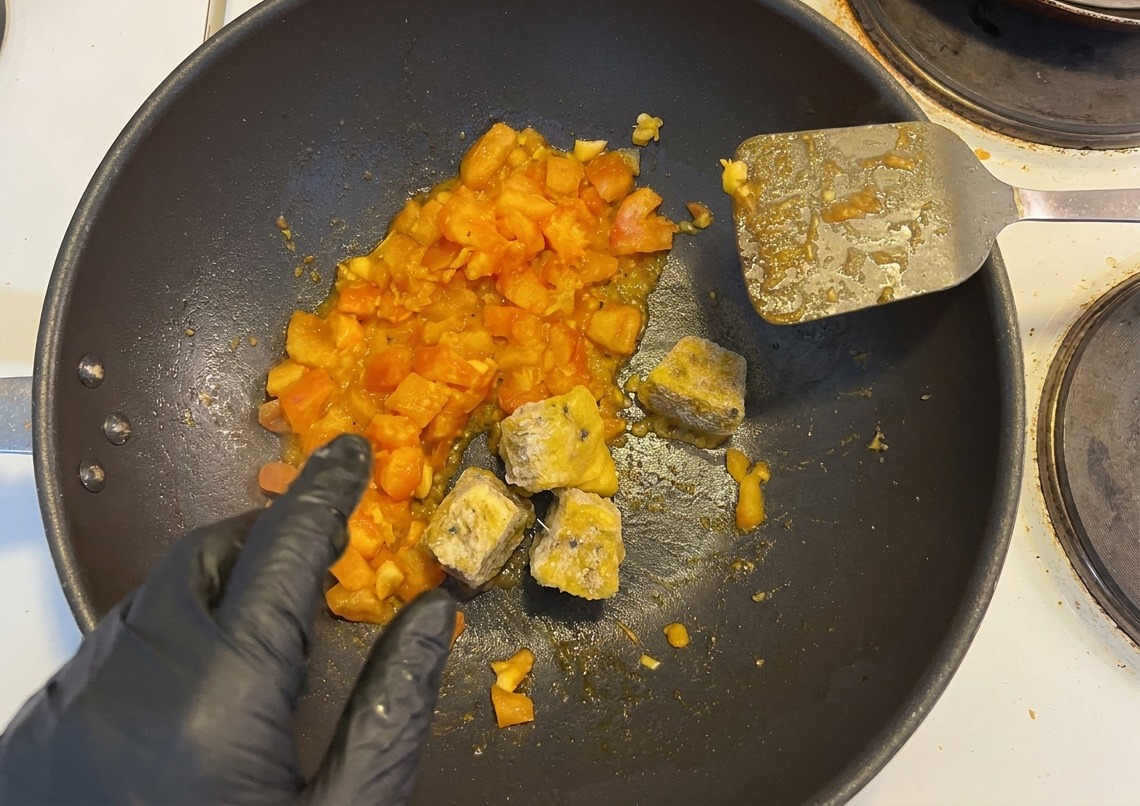 Mrożone kostki pulpy z marakui i posiekane marynowane papryczki dodawane do patelni z podsmażonymi pomidorami przez dłoń w czarnej rękawiczce.