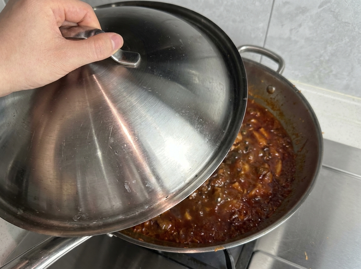 Ręka nakładająca stalową pokrywkę na dużą patelnię typu wok wypełnioną bulgoczącym czerwonym sosem.