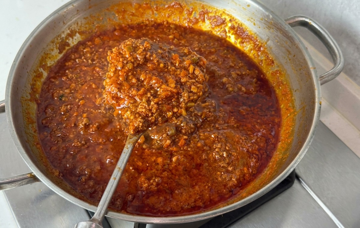 Stalowa patelnia wypełniona gęstym, ciemnoczerwonym sosem bolognese podnoszonym dużą metalową łyżką, by pokazać jego konsystencję.