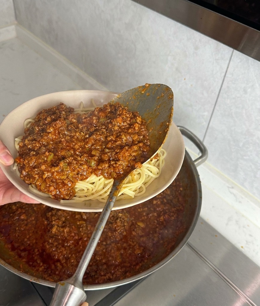 Duża metalowa chochla nakładająca gęsty sos mięsny na porcję ugotowanego makaronu spaghetti w beżowej misce.