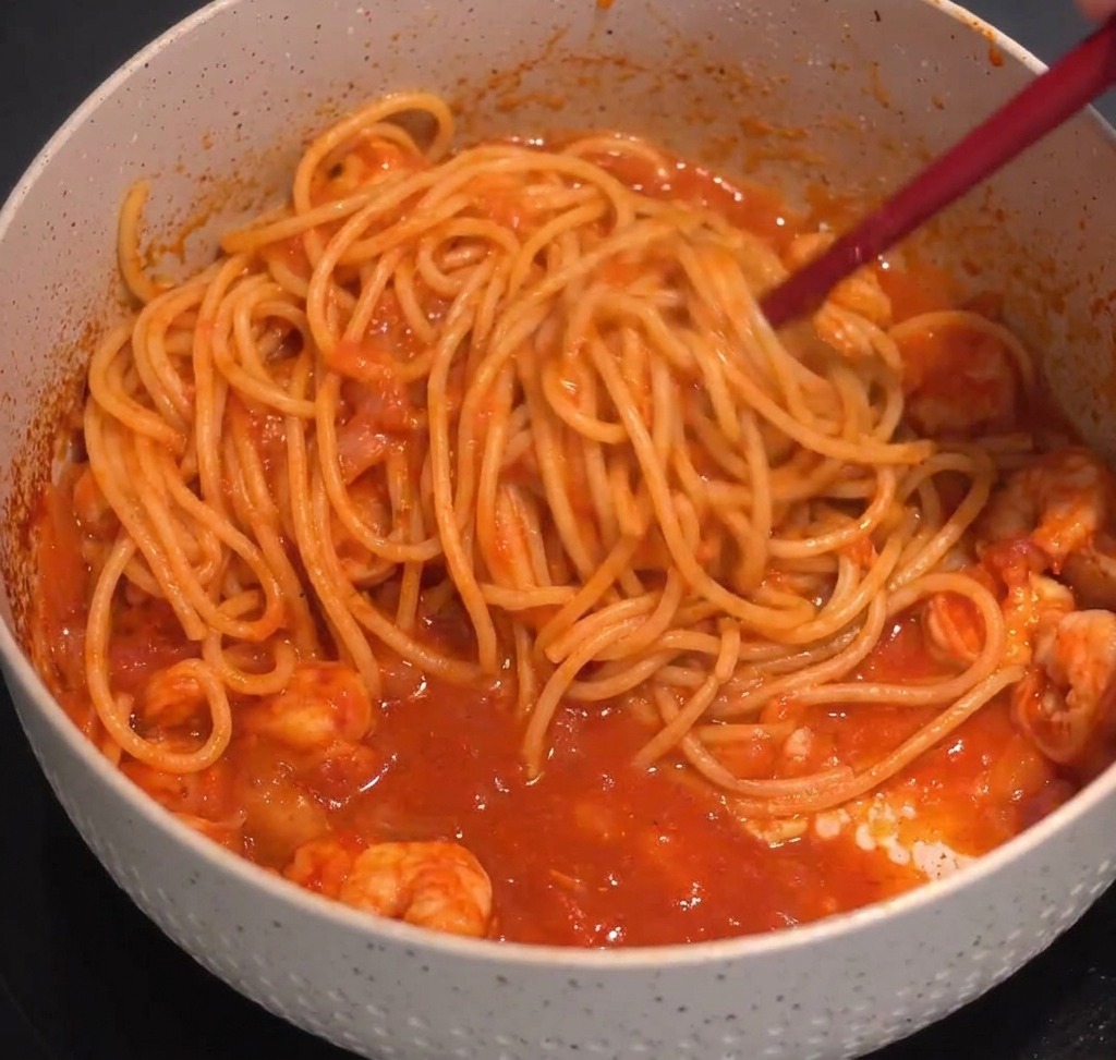 Makaron spaghetti dokładnie mieszany szczypcami z czerwonym sosem na patelni.