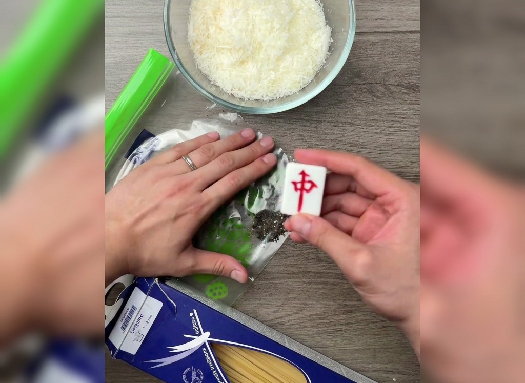 Ręka używająca płaskiej strony kostki mahjong do kruszenia ziaren pieprzu w plastikowej torebce na drewnianym stole.
