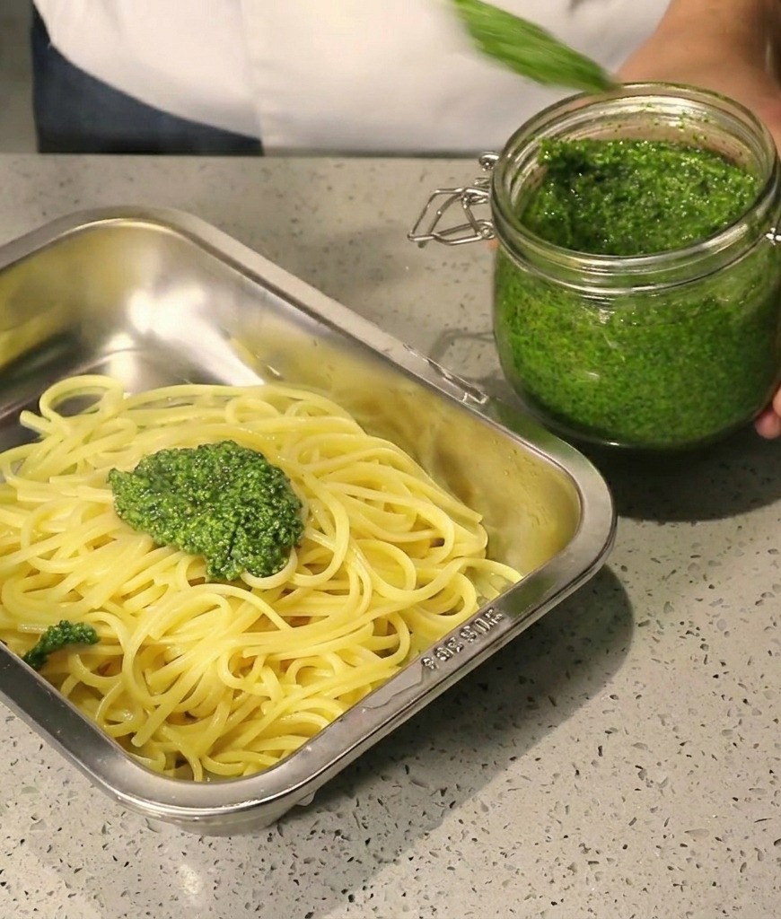 Gęste zielone pesto nakładane łyżką na gorące spaghetti w stalowej tacy.