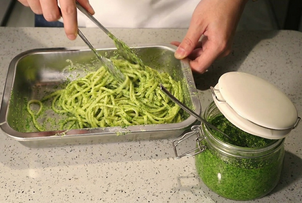 Mieszanie spaghetti z pesto i śmietanką za pomocą szczypiec w metalowej misce.