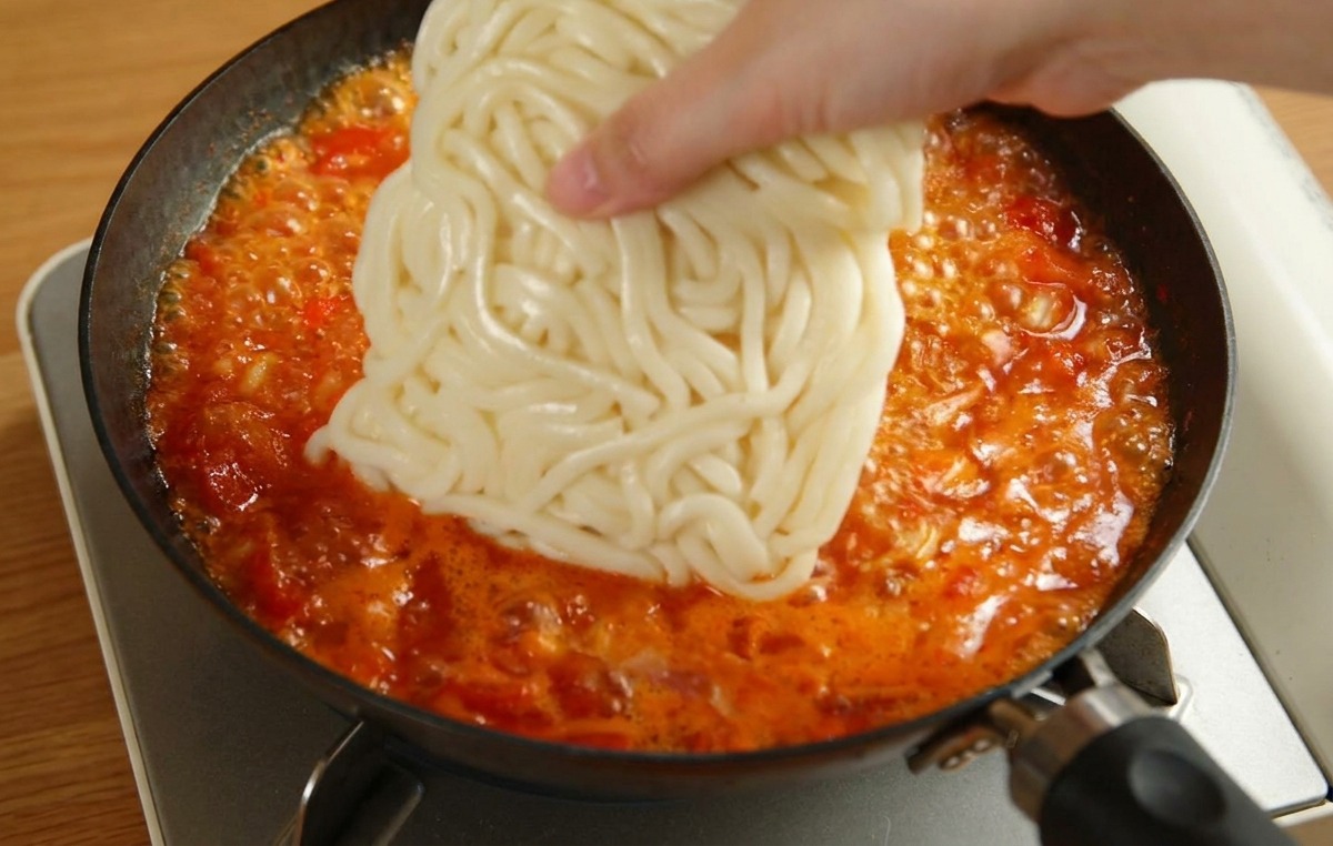 Dłoń kładąca kostkę białego makaronu udon bezpośrednio do gotującego się czerwonego sosu pomidorowego.