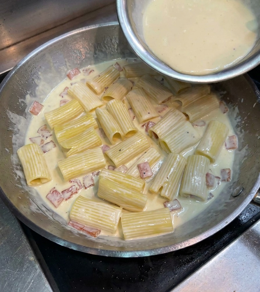 Wlewanie złocistej masy jajecznej z serem na makaron rigatoni na dużej patelni.
