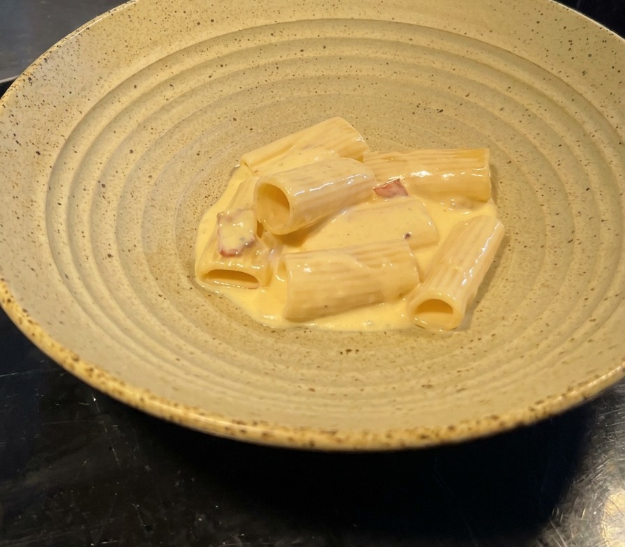 Porcja rigatoni w lśniącym sosie z kawałkami boczku podana w szerokiej ceramicznej misce.