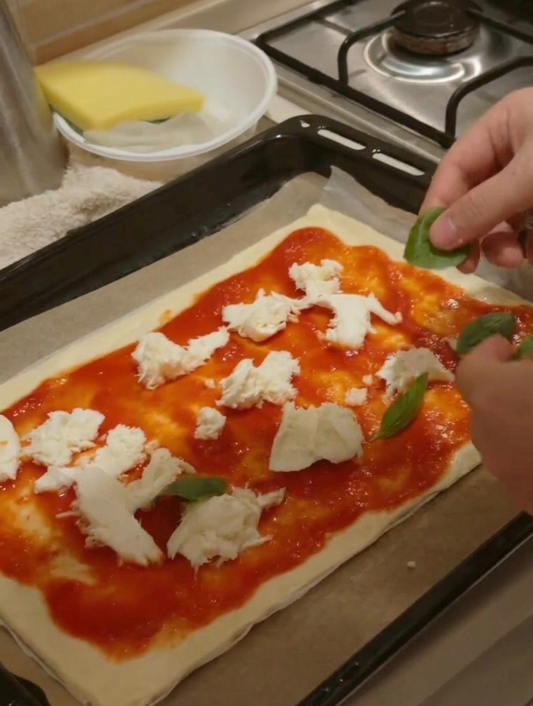 Dłonie kładące świeże liście zielonej bazylii na pizzę z sosem i kawałkami sera.