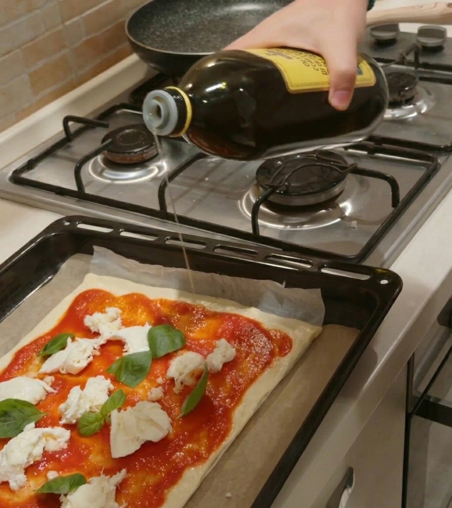 Dłoń polewająca strużką oliwy z dużej szklanej butelki surową pizzę z sosem, mozzarellą i bazylią.
