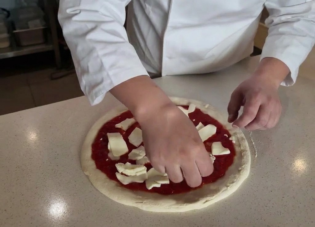 Ręce kucharza układające porwane kawałki białej mozzarelli na sosie pomidorowym.