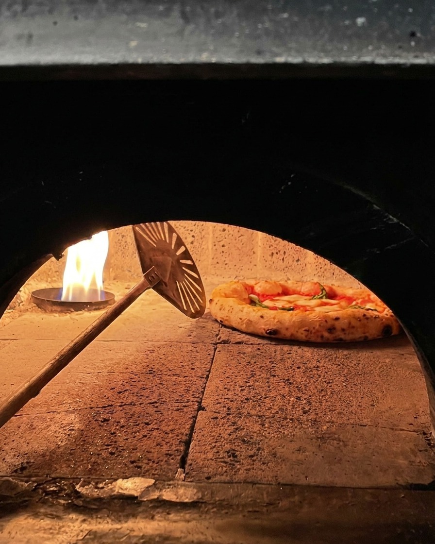 Pizza Margherita pieczona w kamiennym piecu, obracana małą metalową łopatką przy płomieniach.