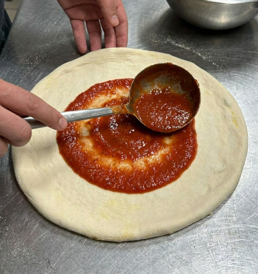 Metalowa chochla rozprowadzająca intensywnie czerwony sos pomidorowy kolistymi ruchami na spodzie pizzy.