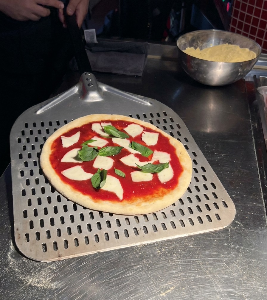 Surowa pizza Margherita z dodatkami spoczywająca na dużej, perforowanej metalowej łopacie do pizzy.