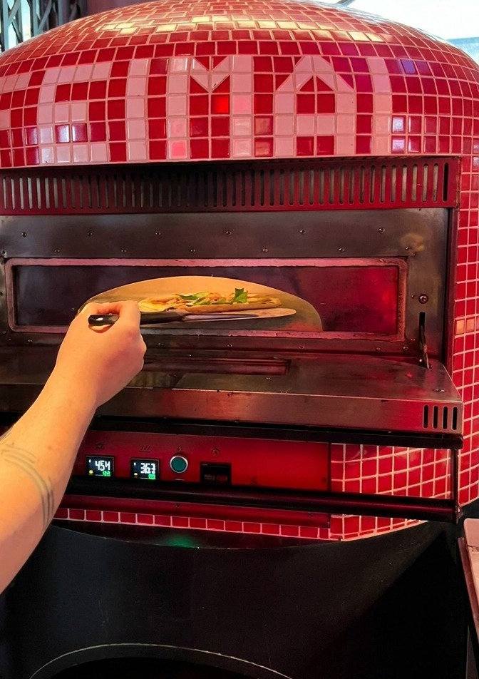 Osoba wsuwająca surową pizzę z metalowej łopaty do rozgrzanego wnętrza tradycyjnego pieca do pizzy.