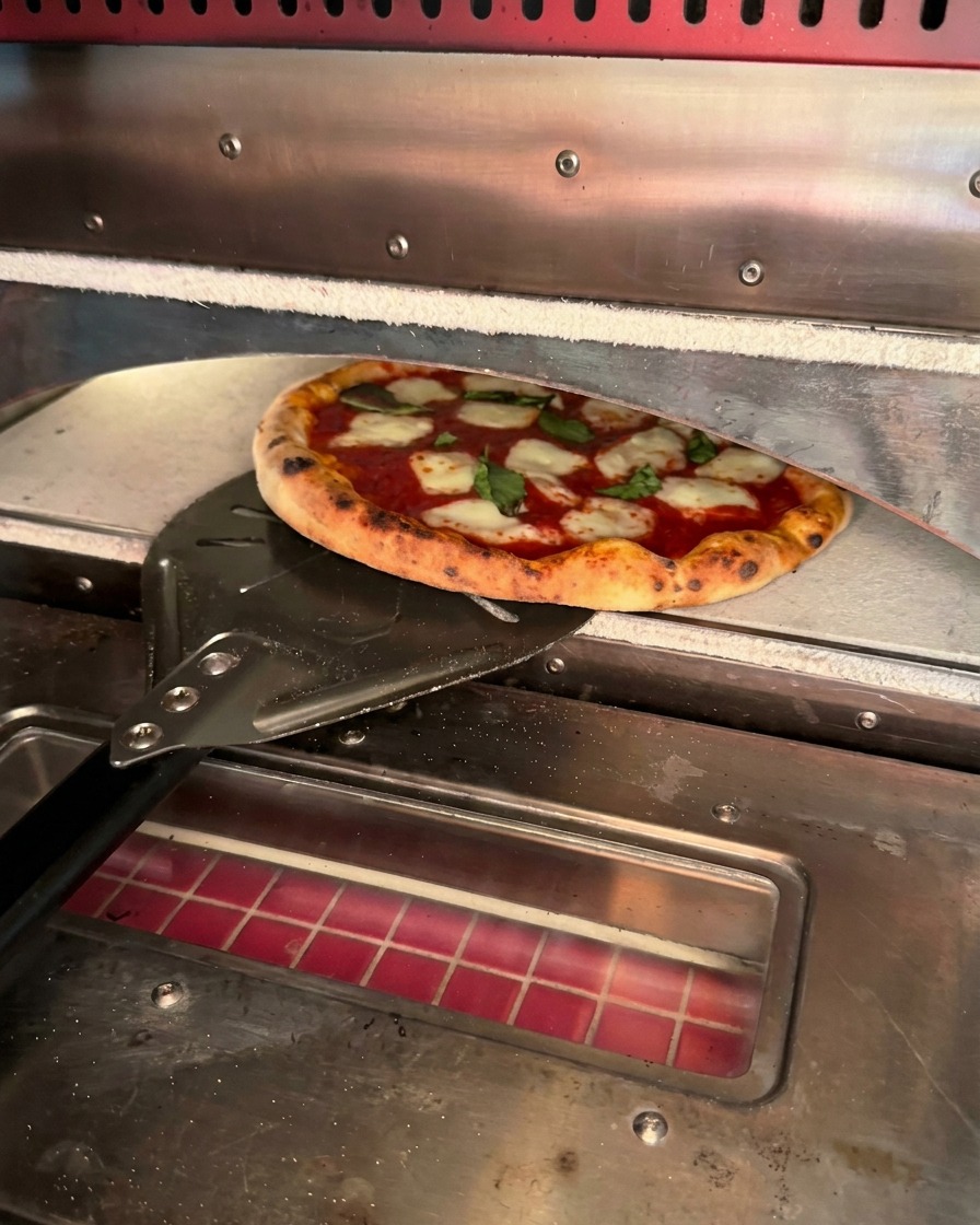 Świeżo upieczona pizza z przypieczonymi brzegami wyjmowana z pieca za pomocą metalowej łopaty.