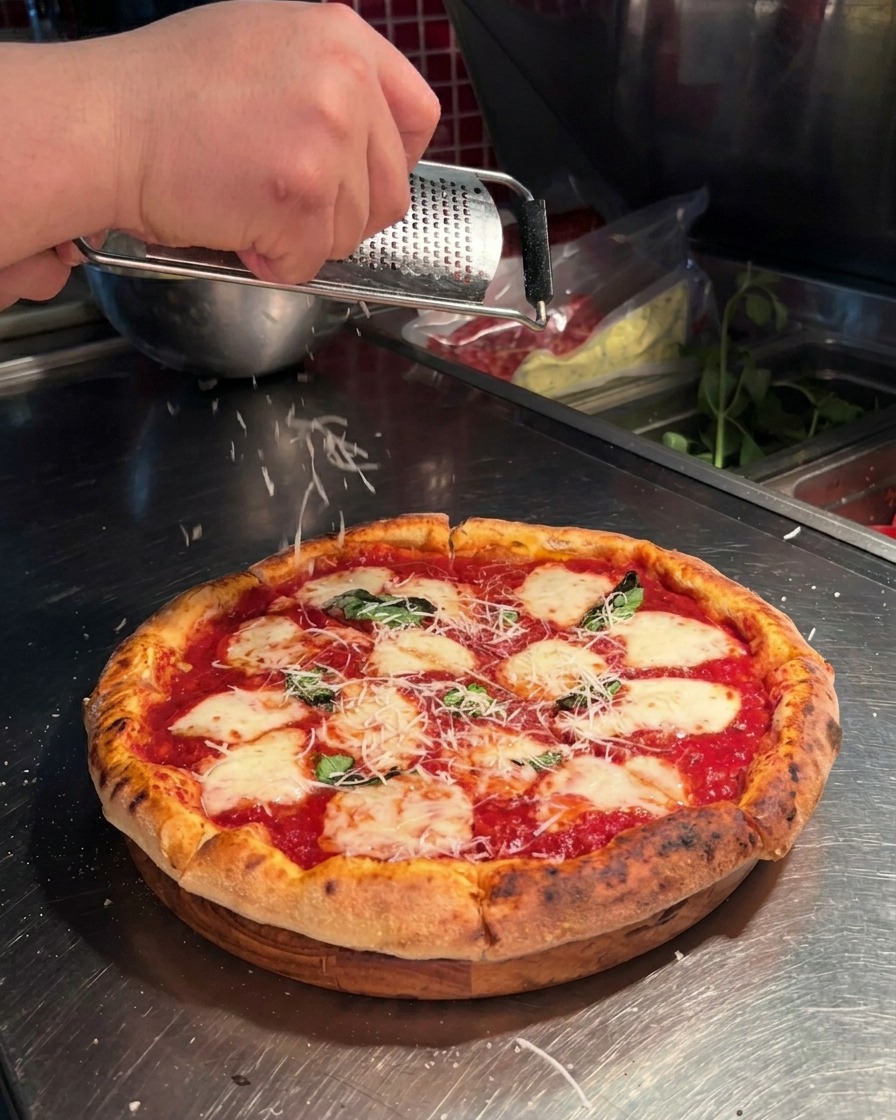 Zbliżenie na dłoń używającą tarki do ścierania świeżego parmezanu nad pizzą w stylu neapolitańskim.