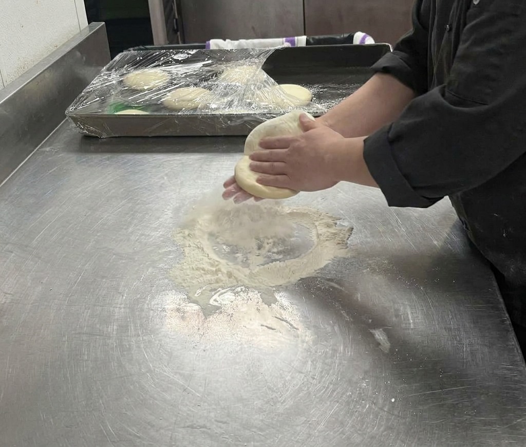 Dłonie rozciągające jasne ciasto na pizzę nad blatem, formując coraz większe koło.