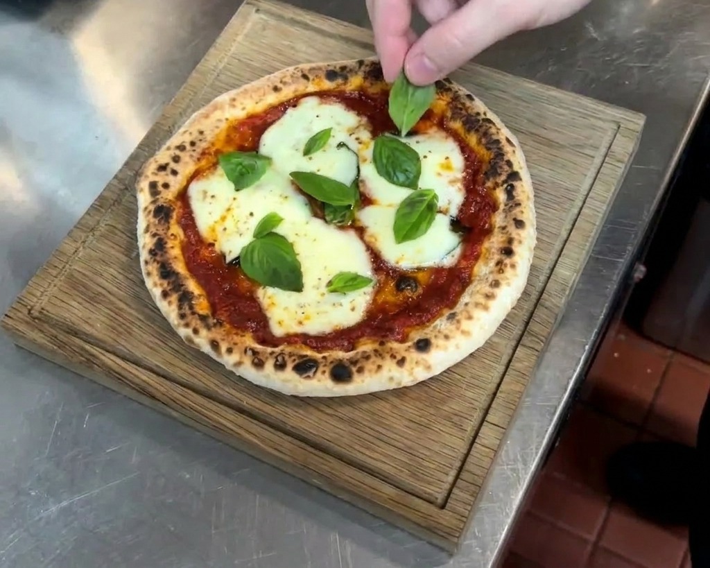 Dłoń układająca świeże liście bazylii na gorącej, upieczonej pizzy z bąblami na brzegach.