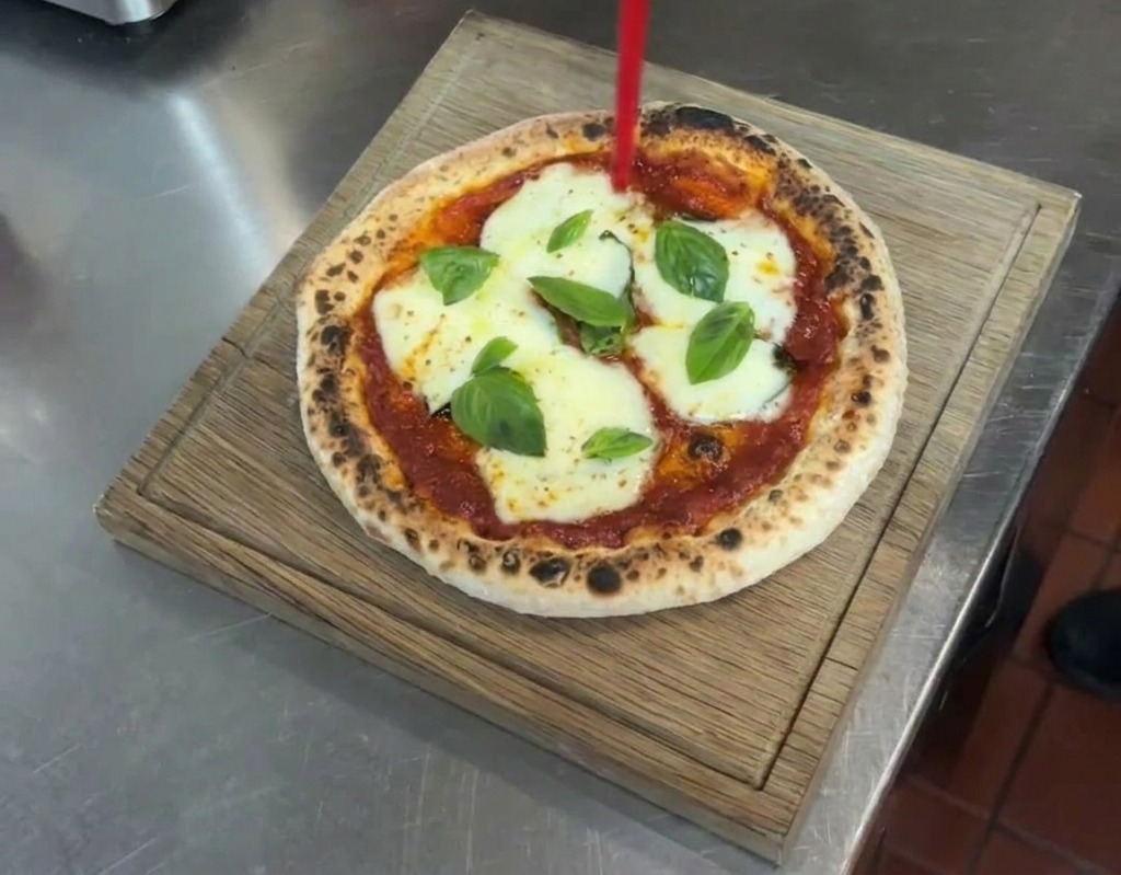 Strumień oliwy extra virgin z dozownika na gotową pizzę Margherita leżącą na drewnianej desce.