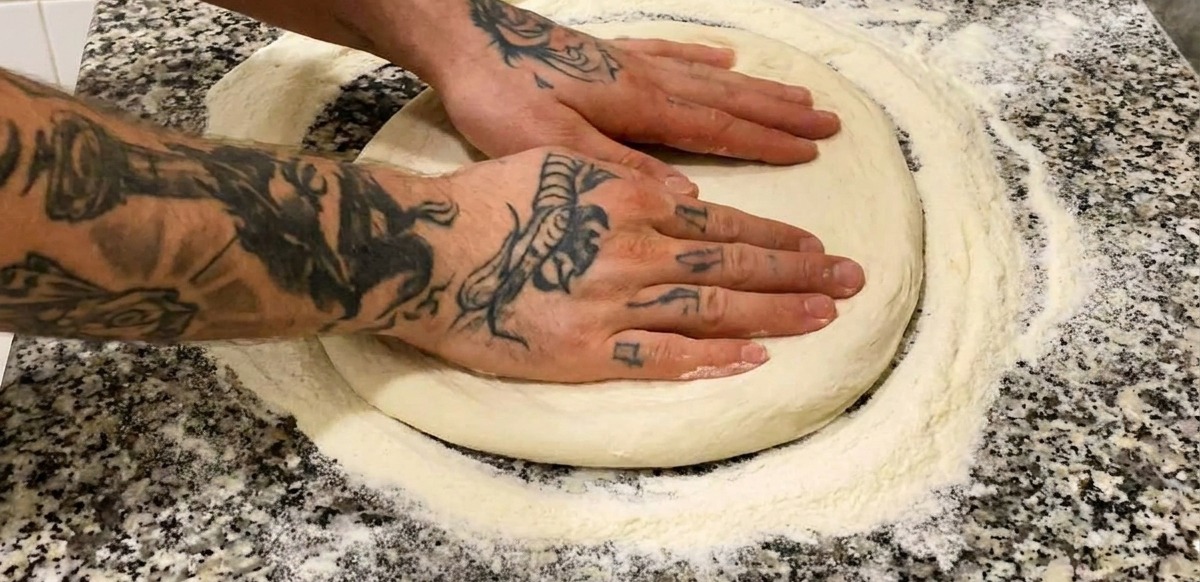Dwie dłonie z tatuażami dociskające okrągły, posypany mąką spód pizzy na szarym marmurowym blacie.