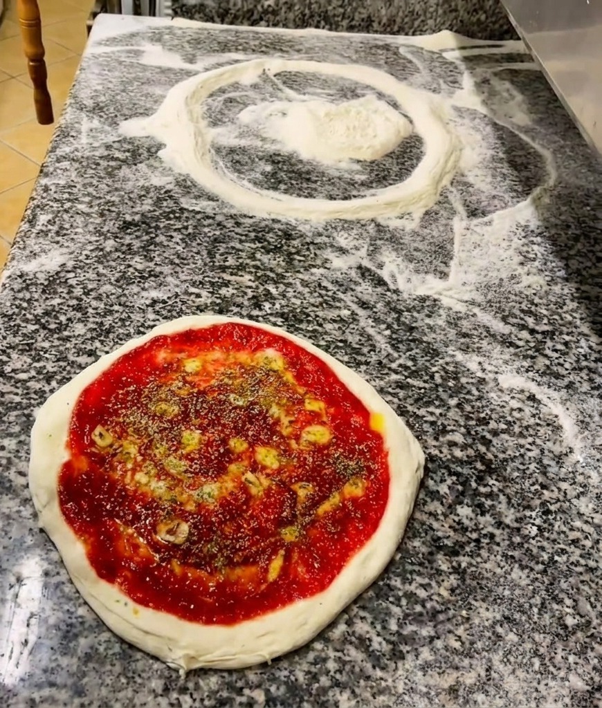 Ręka posypująca zielonymi suszonymi ziołami spód pizzy pokryty sosem i skropiony oliwą.