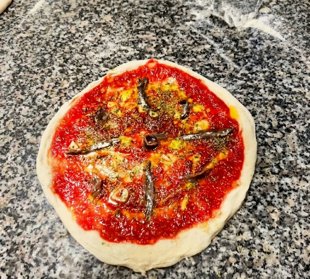 Widok z góry na surową pizzę z sosem, ziołami i kilkoma filetami anchois ułożonymi promieniście.