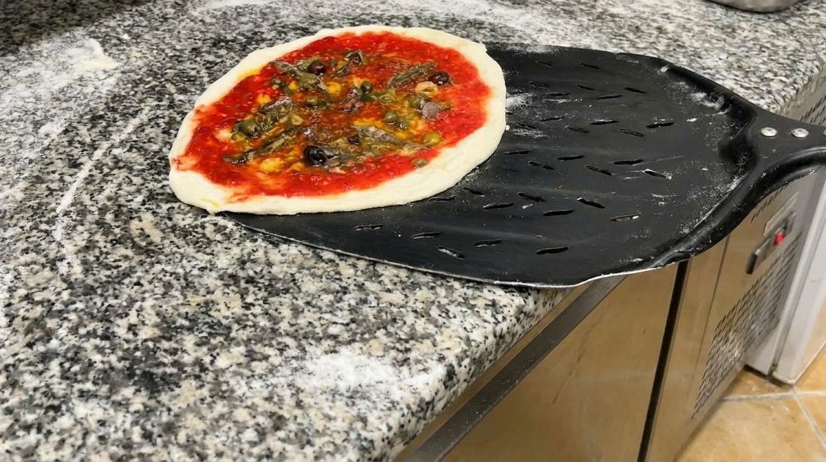 Gotowa do pieczenia pizza z owocami morza na perforowanej czarnej metalowej łopacie na granitowym blacie.