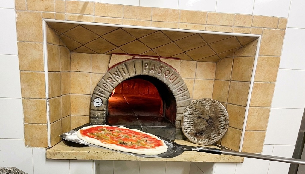 Pizza na łopacie wkładana do tradycyjnego ceglanego pieca z jasnymi pomarańczowymi płomieniami.